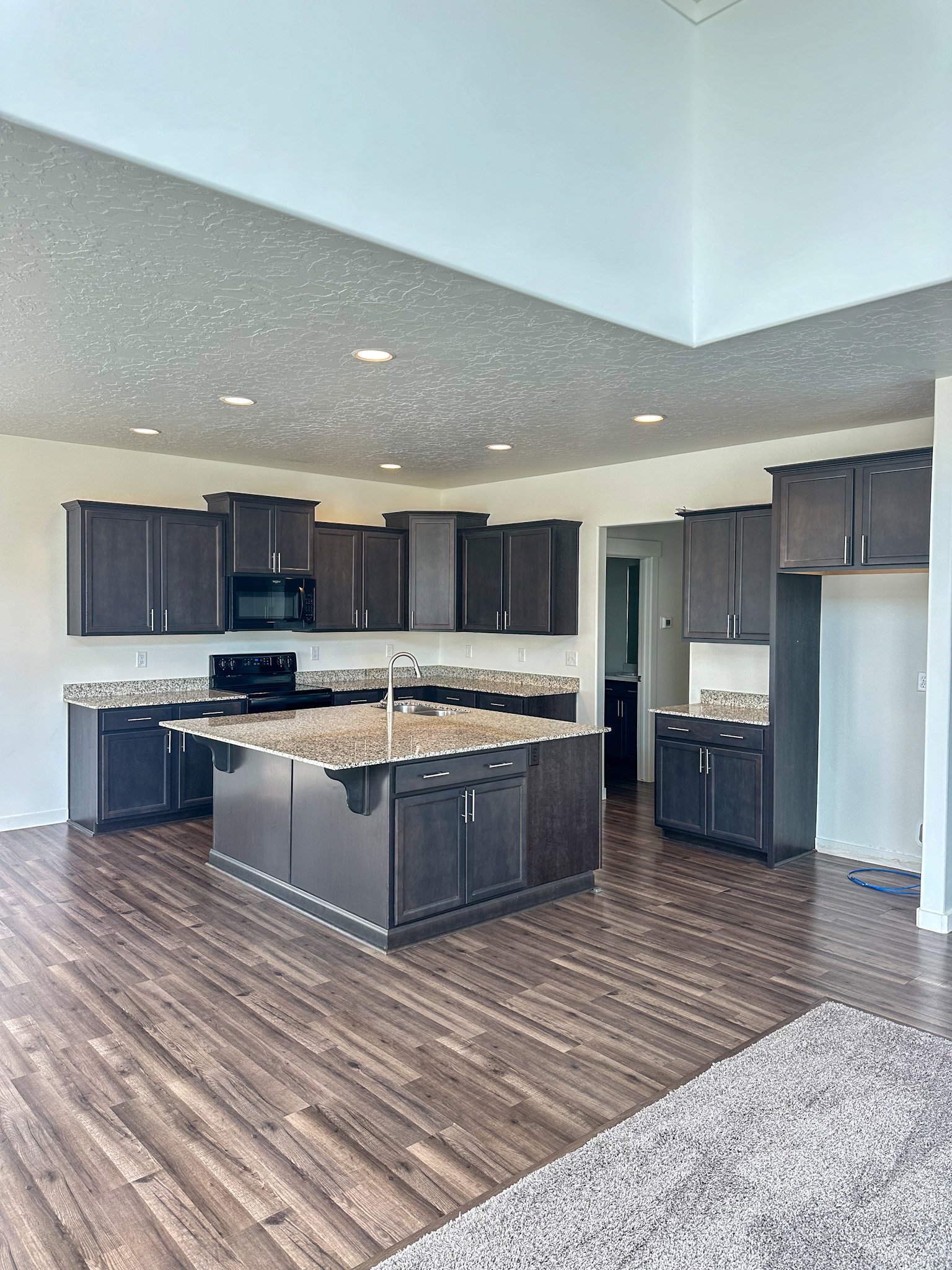 Kitchen featuring dark wood-style flooring, a textured ceiling, a kitchen island with sink, black appliances, and light stone counters