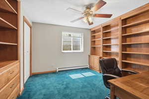 Unfurnished office with built in shelves, a baseboard heating unit, a ceiling fan, and dark colored carpet