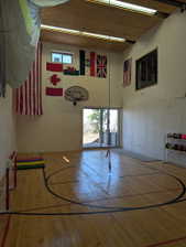 Spacious recreational area featuring wood-finish flooring with court markings, a basketball hoop, and high ceilings with exposed beams