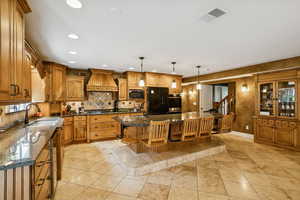 Kitchen with brown cabinetry, a kitchen island with sink, pendant lighting, dark stone counters, and recessed lighting