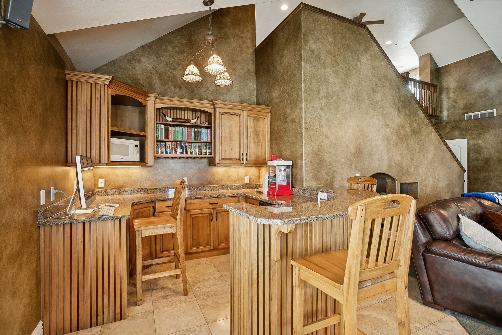 Bar area with high vaulted ceiling, hanging light fixtures, white microwave, open shelves, and brown cabinets