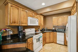 Kitchen featuring white appliances, light tile patterned floors, dark stone counters, brown cabinetry, and recessed lighting