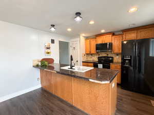 Kitchen featuring a central island with a white basin sink and matte black faucet, dark stone countertops, wood-finish cabinetry, black appliances, and wood-finish flooring