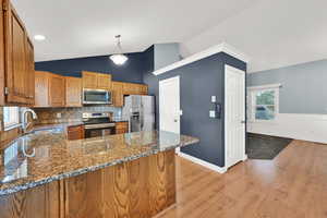 Kitchen featuring vaulted ceiling, dark stone countertops, stainless steel appliances, wood finish cabinets, and decorative light fixtures