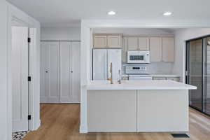 Kitchen with white appliances, light wood-type flooring, a center island with sink, recessed lighting, and light stone countertops