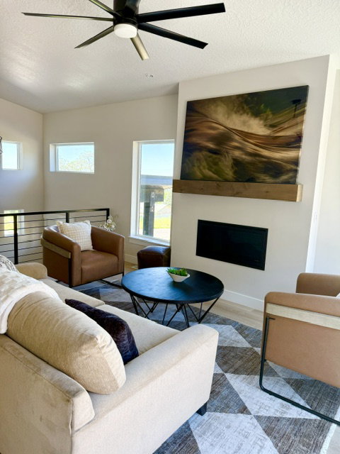 Living area featuring a modern black ceiling fan, multiple windows providing natural light, a fireplace with a wood mantel, and wood-finish flooring with an area rug