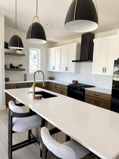 Kitchen featuring a long white countertop island with integrated sink, two-tone cabinetry with wood-finish base cabinets and white upper cabinets, a black range hood, and three black pendant lights