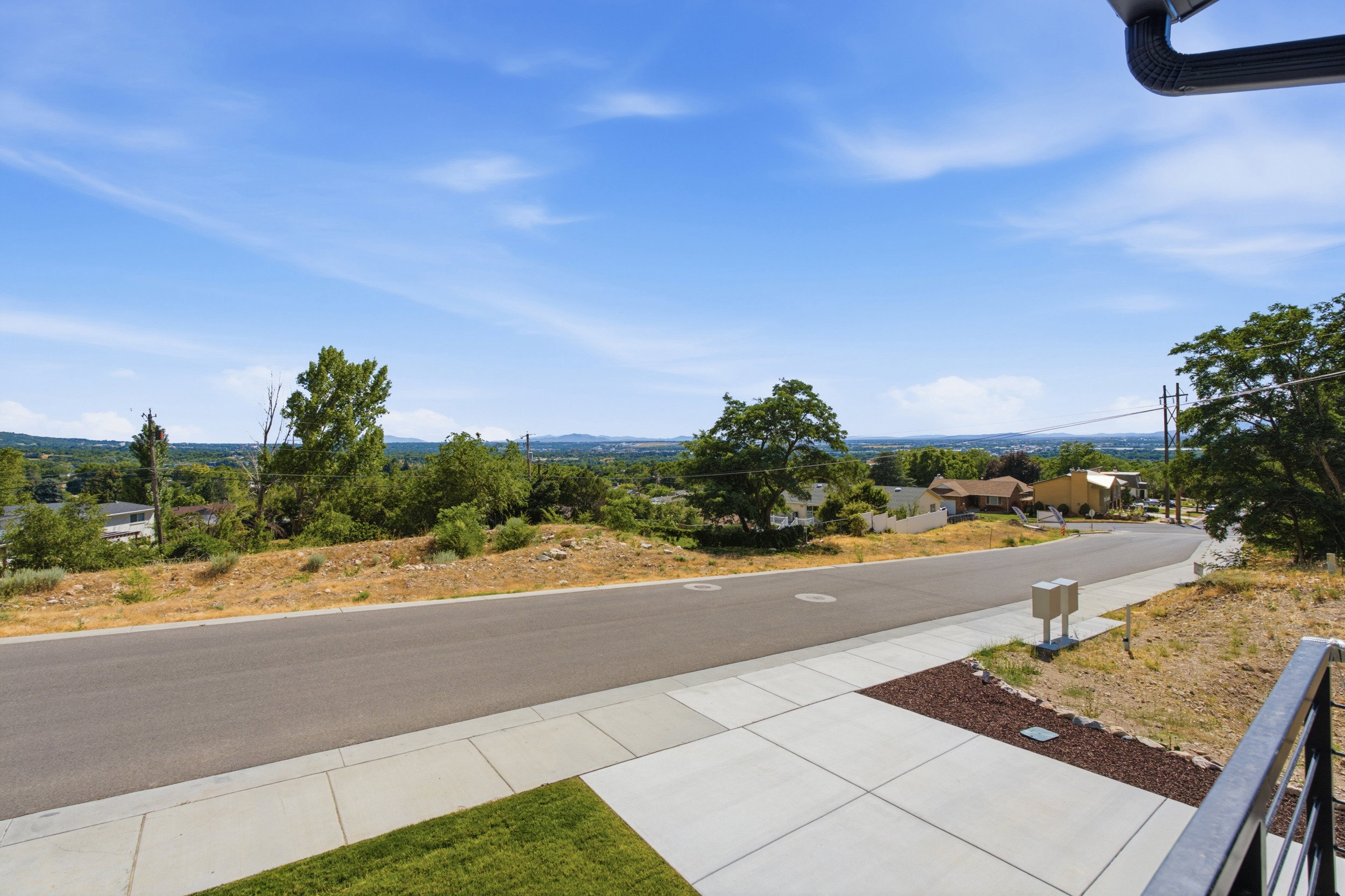 Elevated property with a paved street frontage, concrete sidewalk, and a distant cityscape horizon