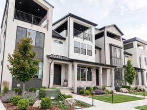 Modern townhome exterior featuring an open-air balcony, a covered porch entry, stacked stone accents, dark frame windows, and manicured landscaping