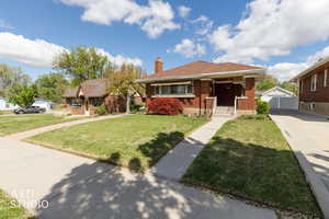 Bungalow-style house featuring a front yard, brick siding, a chimney, a garage, and an outbuilding
