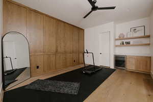 Exercise/flex area with beverage cooler, light wood-type flooring, wooden walls, a ceiling fan, and a bar