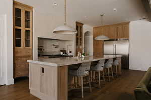 Kitchen featuring glass insert cabinets, stainless steel built in refrigerator, backsplash, dark wood-type flooring, and a large island with sink