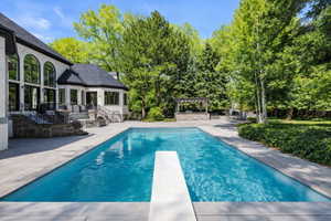 Saline swimming pool with a patio area, a diving board, view of wooded area, and a pergola with outdoor kitchen.