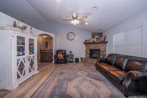 Living area featuring lofted ceiling, a ceiling fan, wood finished floors, a stone fireplace, and arched walkways