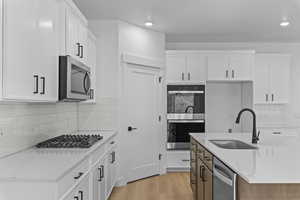 Dual tone kitchen featuring light stone counters, stainless steel appliances, backsplash, light wood finished floors, and an island with sink