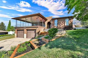 View of front of property featuring brick siding, driveway, a balcony, an attached garage, and a garden