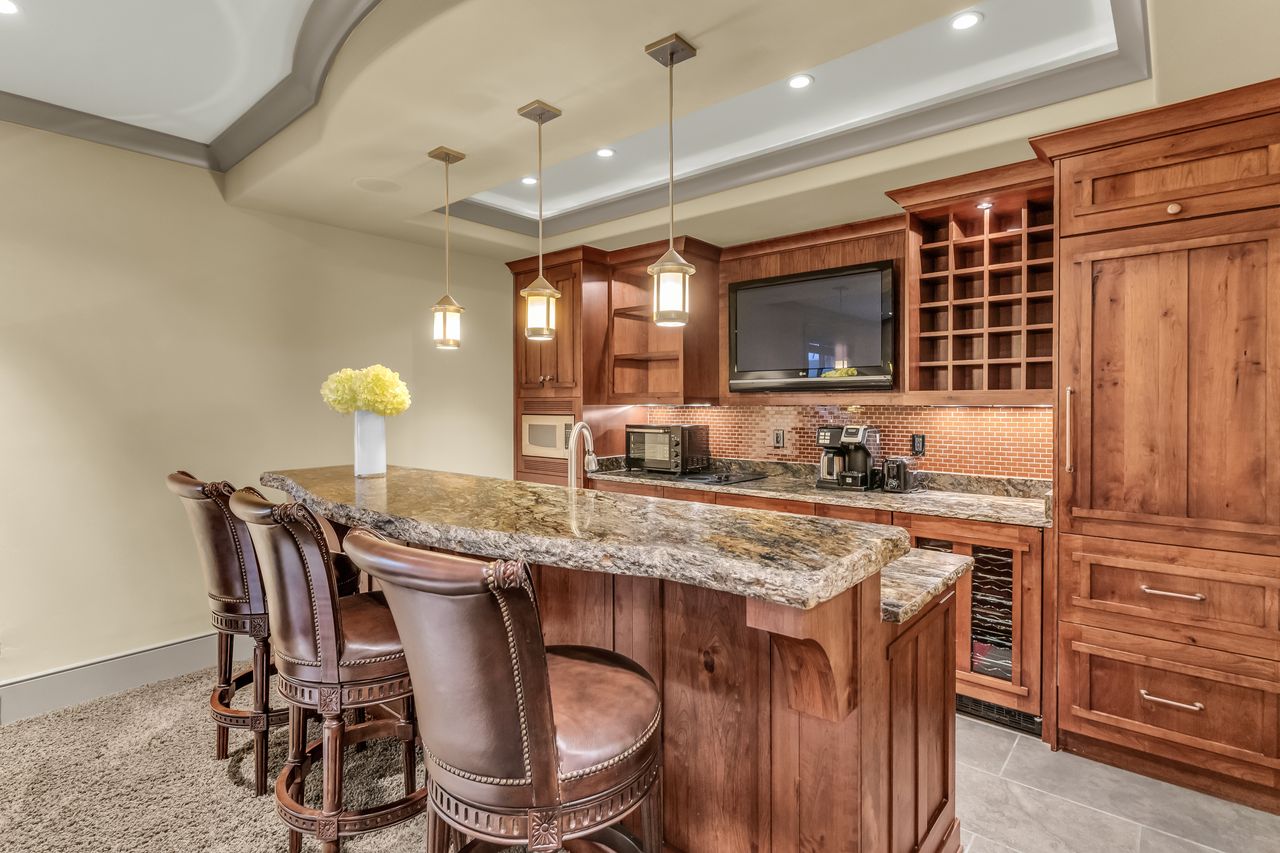 Indoor wet bar with wood finish cabinets, a raised ceiling, light stone countertops, decorative light fixtures, and backsplash