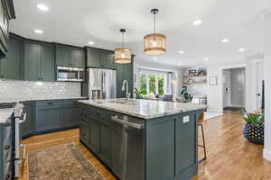 Kitchen with light stone counters, stainless steel appliances, a kitchen island with sink, green cabinetry, and light wood finished floors