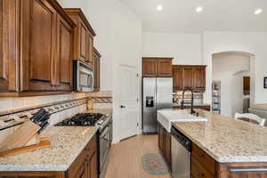 Kitchen featuring stainless steel appliances, light stone countertops, a kitchen island with sink, arched walkways, and light wood-style flooring