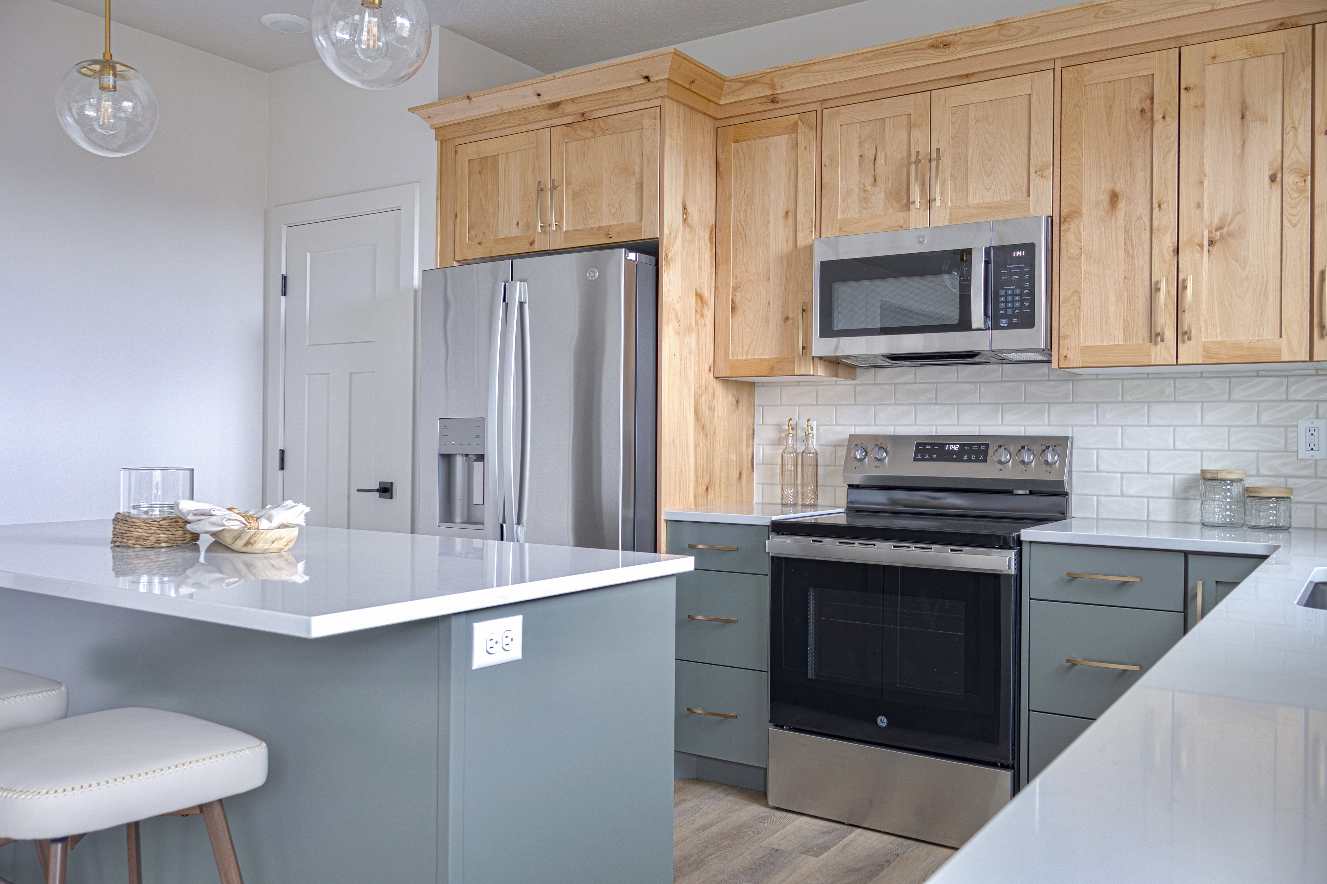 Kitchen featuring a two-tone cabinet design with natural wood and muted green finishes, stainless steel appliances, white subway tile backsplash, and light-toned countertops