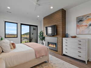 Bedroom featuring access to outside, a glass covered fireplace, wood finished floors, a ceiling fan, and recessed lighting