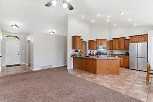 Kitchen with wood finish cabinetry, stainless steel appliances, light tile patterned floors, light colored carpet, and vaulted ceiling