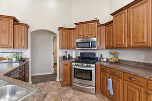Kitchen featuring stainless steel appliances, wood finish cabinets, dark countertops, arched walkways, and a high ceiling