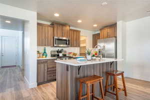 Kitchen with a breakfast bar area, wood finish cabinetry, light wood finished floors, stainless steel appliances, and an island with sink
