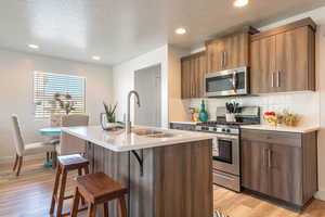 Kitchen featuring stainless steel appliances, a kitchen bar, light stone counters, a center island with sink, and wood finish cabinets