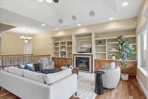 Living room featuring wood finished floors, a tile fireplace, built in features, a ceiling fan, and a tray ceiling