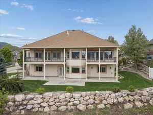 Back of house featuring a patio, a lawn, stucco siding, roof with shingles, and a balcony