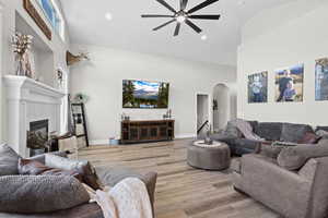 Living room with a high ceiling, arched walkways, light wood-type flooring, ceiling fan, and a glass covered fireplace