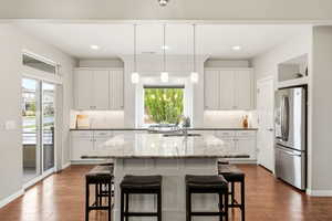 Kitchen featuring stainless steel refrigerator with ice dispenser, white cabinets, a kitchen island with sink, light stone countertops, and decorative light fixtures