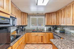 Kitchen featuring black appliances, dark stone countertops, light tile patterned floors, tasteful backsplash, and wood finish cabinetry