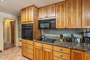 Kitchen with black appliances, light tile patterned floors, dark stone countertops, and backsplash