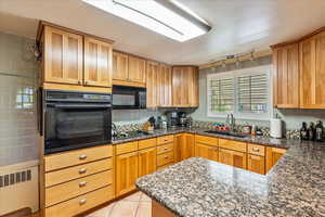 Kitchen with dark stone counters, black appliances, light tile patterned flooring, radiator, and wood finish cabinetry