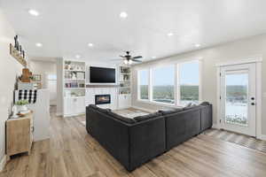 Living room with light wood-style flooring, a glass covered fireplace, a ceiling fan, recessed lighting, and built in shelves