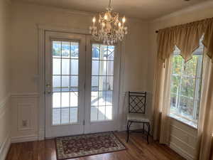 Doorway to outside with a decorative wall, wood finished floors, suspended lighting, wainscoting, and crown molding