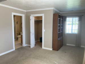 Unfurnished bedroom featuring light colored carpet, ornamental molding, a textured ceiling, a walk in closet, and ensuite bathroom