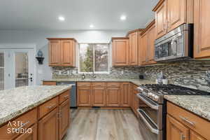 Kitchen with stainless steel appliances, wood finish cabinets, light wood-type flooring, light stone countertops, and backsplash