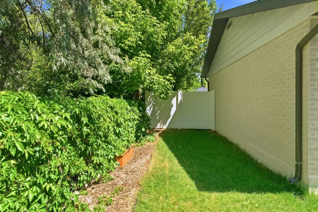 Side yard featuring a white privacy fence, mature green foliage, and a raised garden bed with wood borders