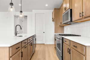 Kitchen featuring stainless steel appliances, light wood-type flooring, a kitchen island with sink, wood finish cabinets, and light stone counters