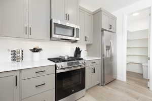 Kitchen featuring gray cabinets, stainless steel appliances, light wood-style floors, decorative backsplash, and light stone counters