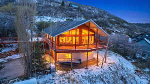 Snow covered back of property featuring a deck with mountain view, a patio area, and a shingled roof
