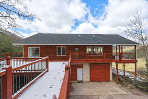 Snow covered back of property featuring a shingled roof, a garage, a wooden deck, and driveway