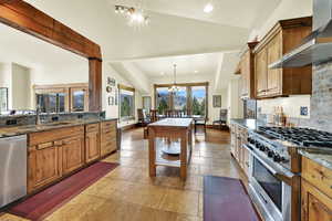 Kitchen featuring stainless steel appliances, wood finish cabinetry, stone tile flooring, dark stone counters, and vaulted ceiling