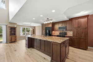 Kitchen featuring a raised ceiling, light wood-type flooring, light stone countertops, black appliances, and a large island with sink
