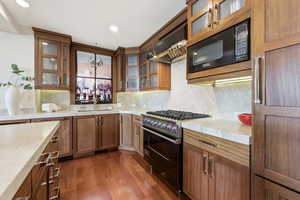 Kitchen with glass insert cabinets, light countertops, black appliances, wood finish cabinetry, and dark wood-style floors