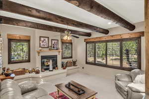 Living room with a glass covered fireplace, carpet flooring, ceiling fan, and beam ceiling