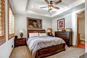 Bedroom with light colored carpet, a ceiling fan, a tray ceiling, ensuite bathroom, and recessed lighting